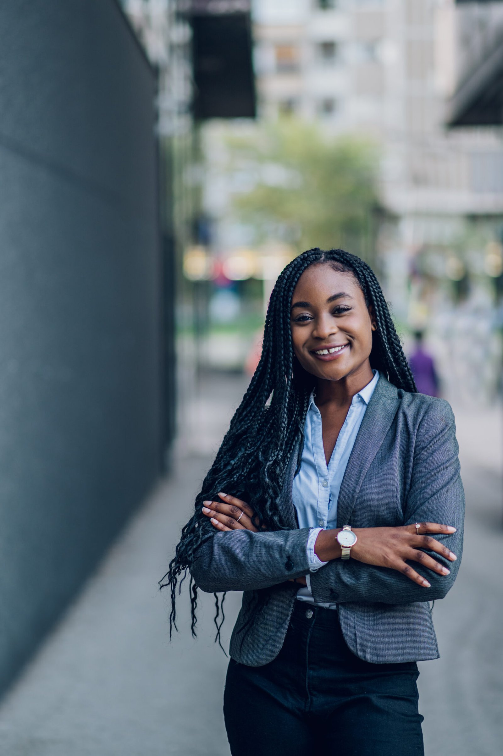 Portrait of a confident african american business woman wearing elegant suit standing at the street with cheerful face and arms crossed in front of the office building. Looking into the camera.