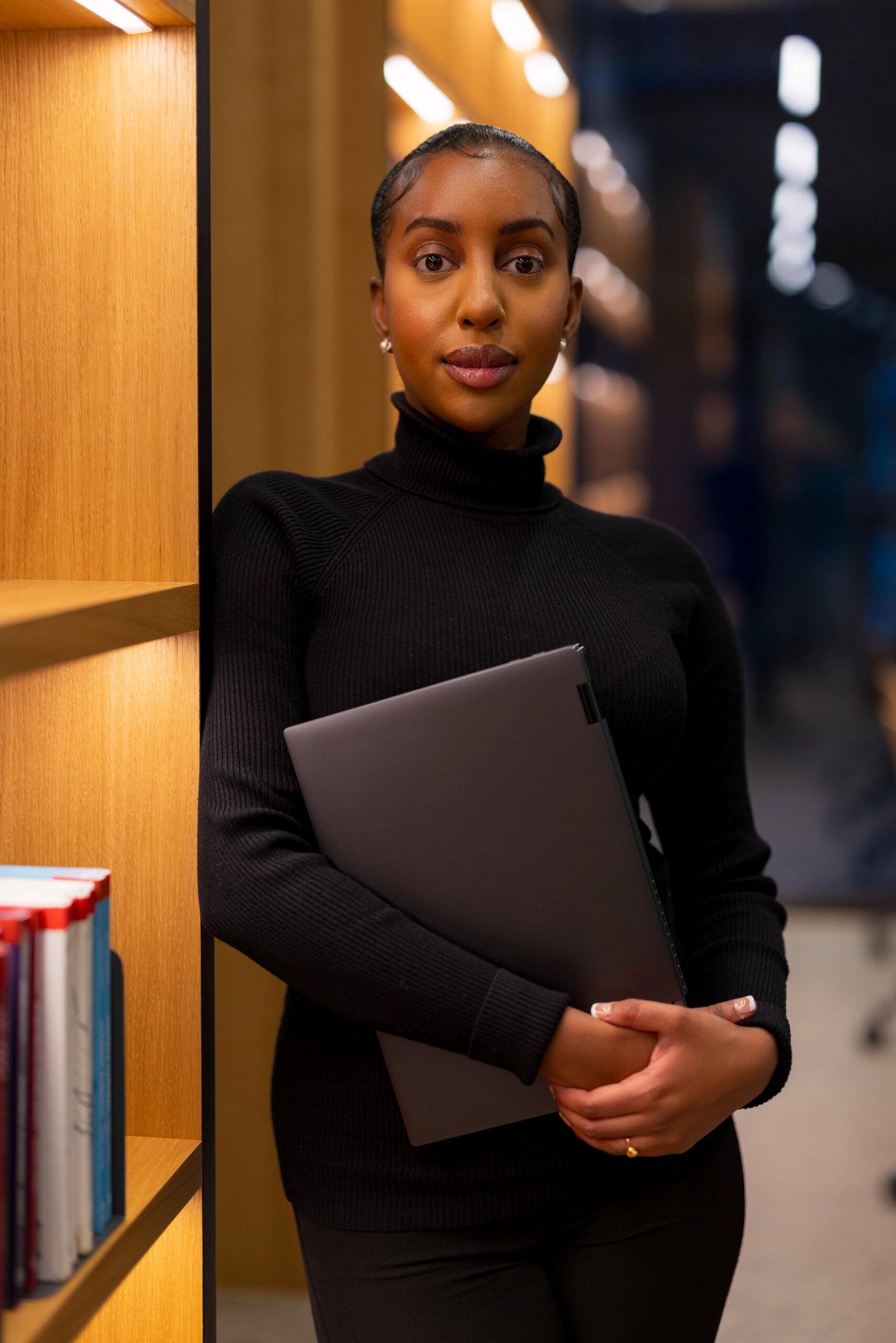 A young student standing by a bookshelf in a library, holding a laptop for her studies. The setting is illuminated with soft, warm lighting, providing a quiet and concentrated atmosphere perfect for study or research in an academic or professional setting.